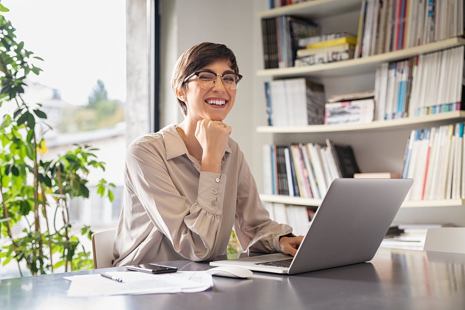 Person smiling at a laptop in a bright home office with bookshelves and a potted plant