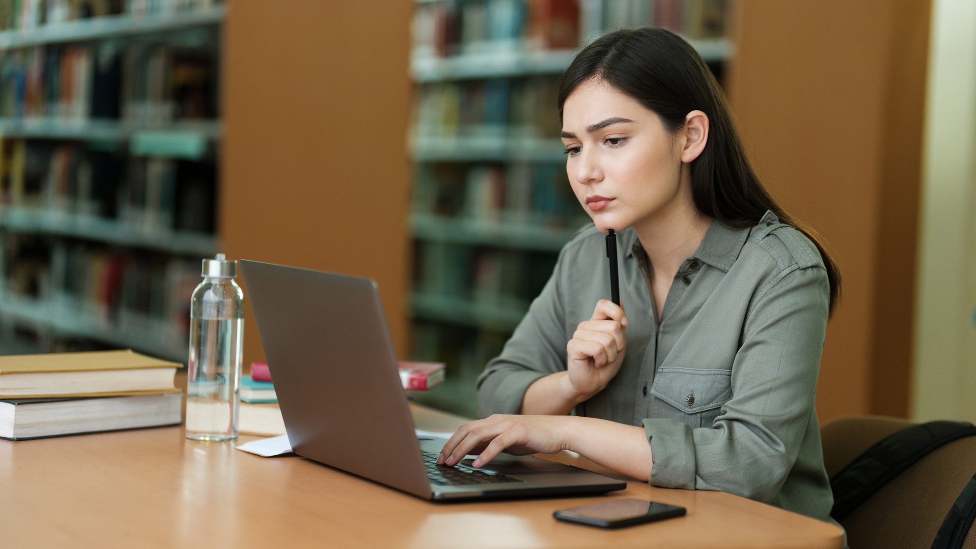 Woman working on a laptop at a library table with books and a water bottle nearby
