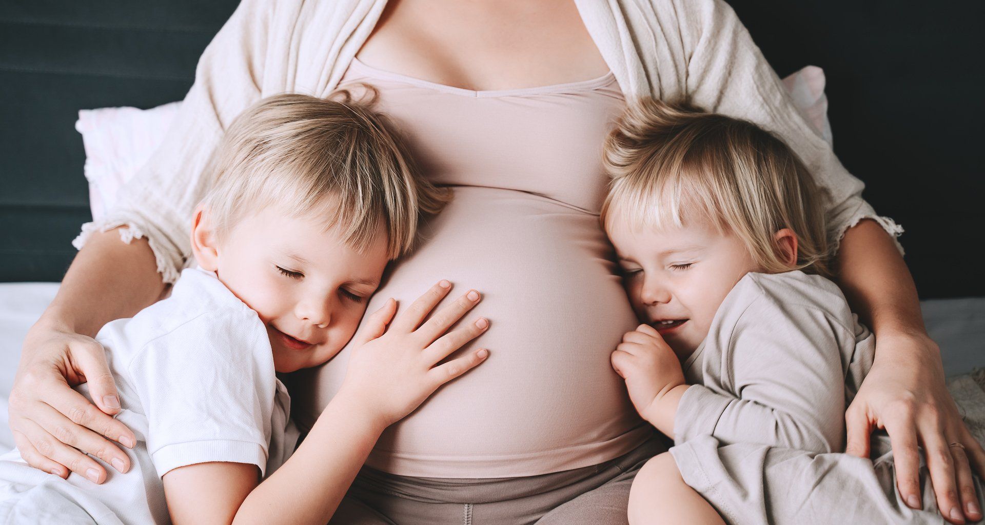 Pregnant person with two sleeping children leaning on their belly on a bed