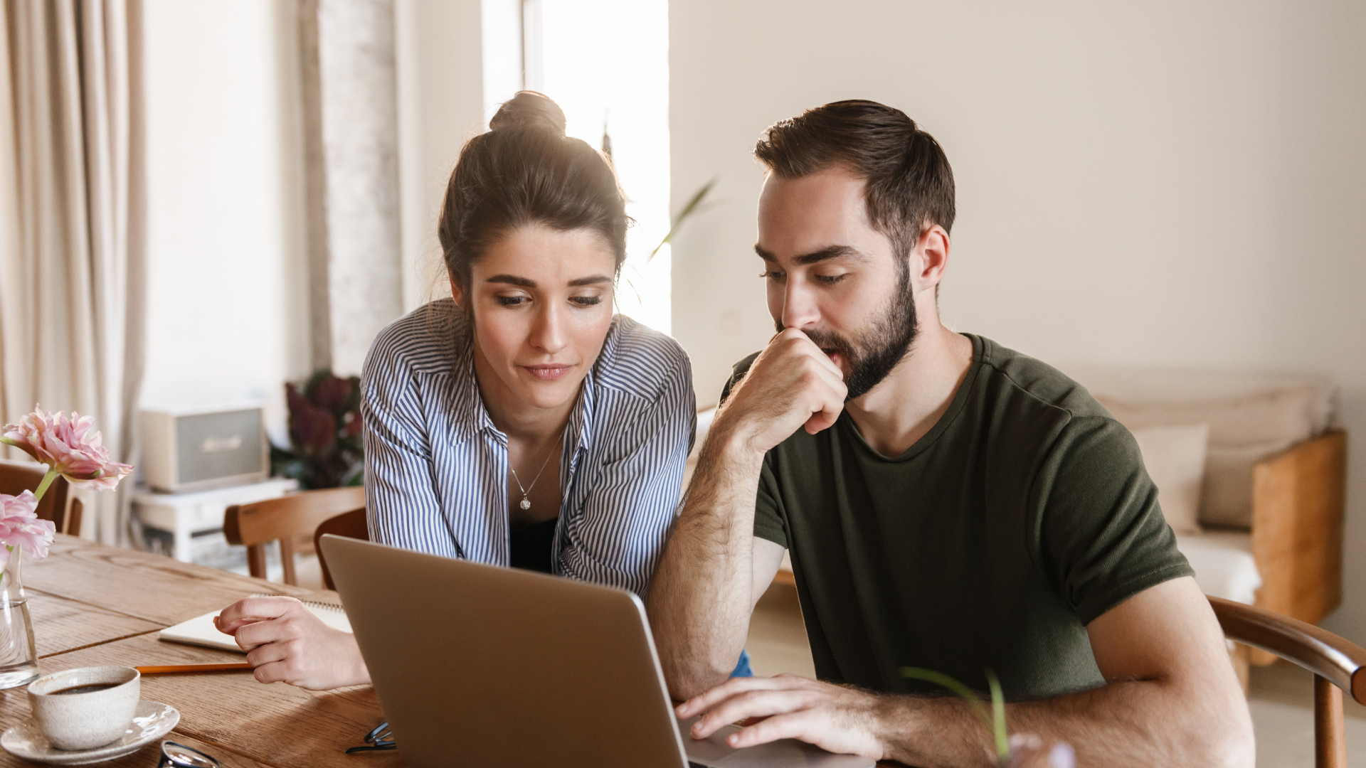 Two people leaning over a laptop at a table, collaborating in a bright home office.