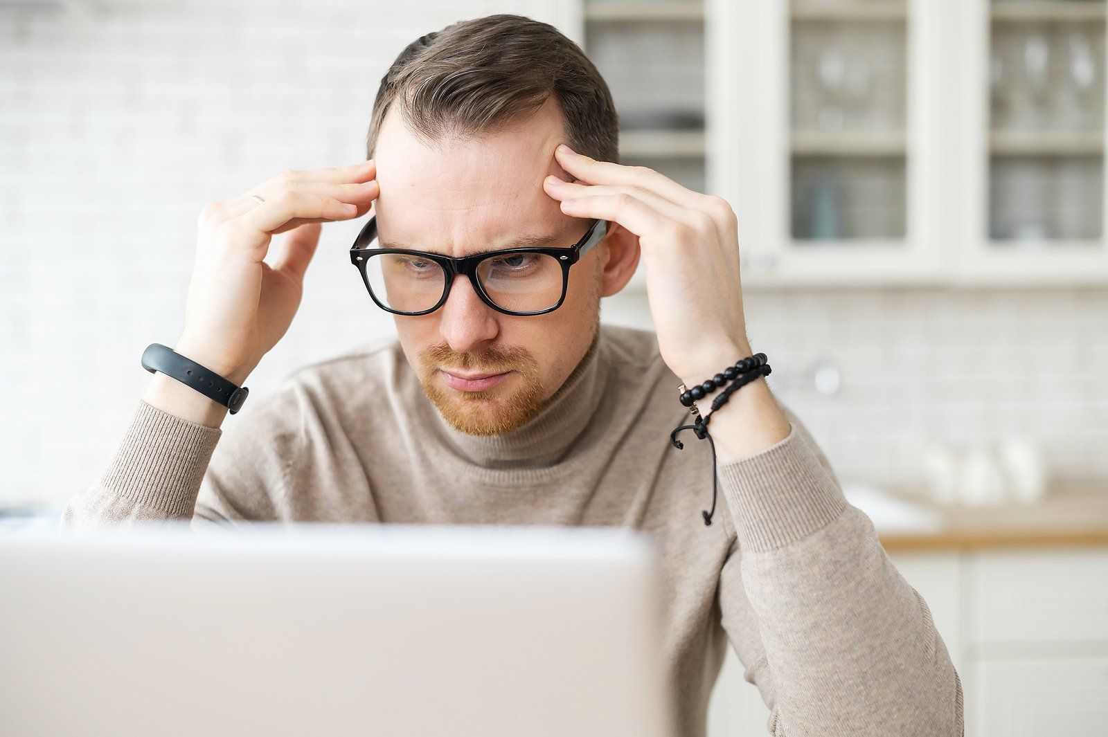Person wearing glasses, holding their temples in frustration while looking at a laptop.