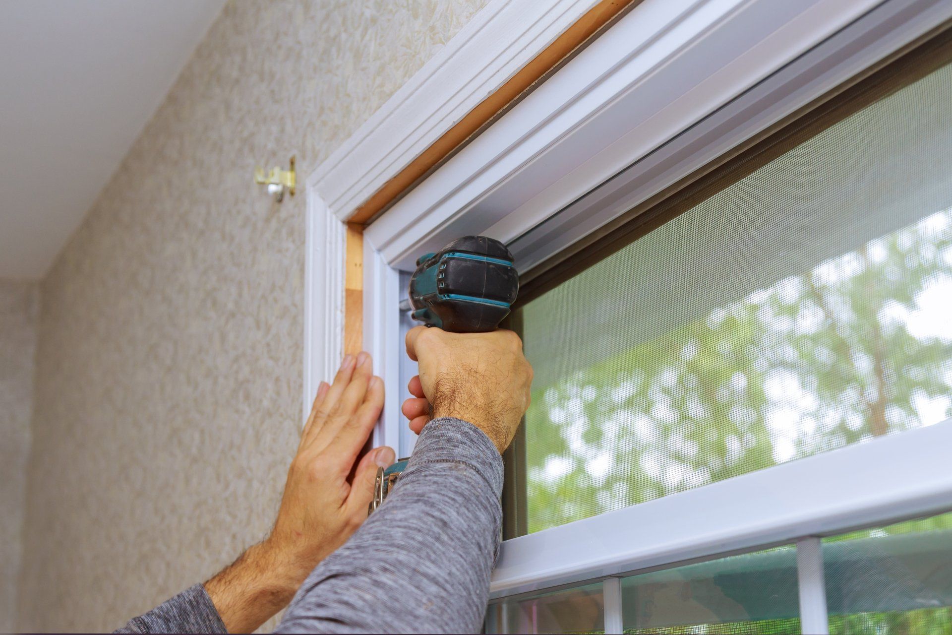 Hands use a drill near the top of a window frame indoors.