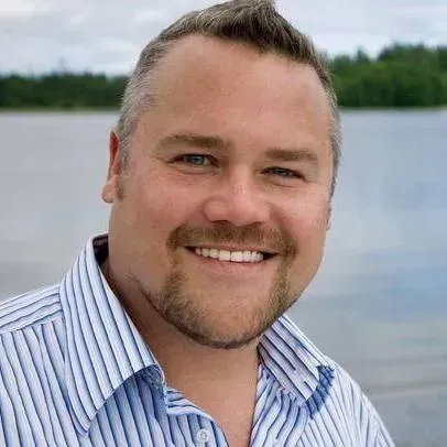 Smiling man in a blue striped shirt standing by a lake with trees in the background