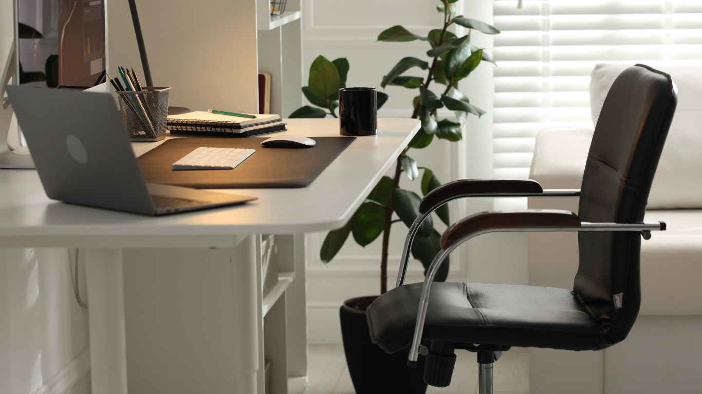 Home office desk with laptop, notebook, coffee mug, plant, and a black chair by a window