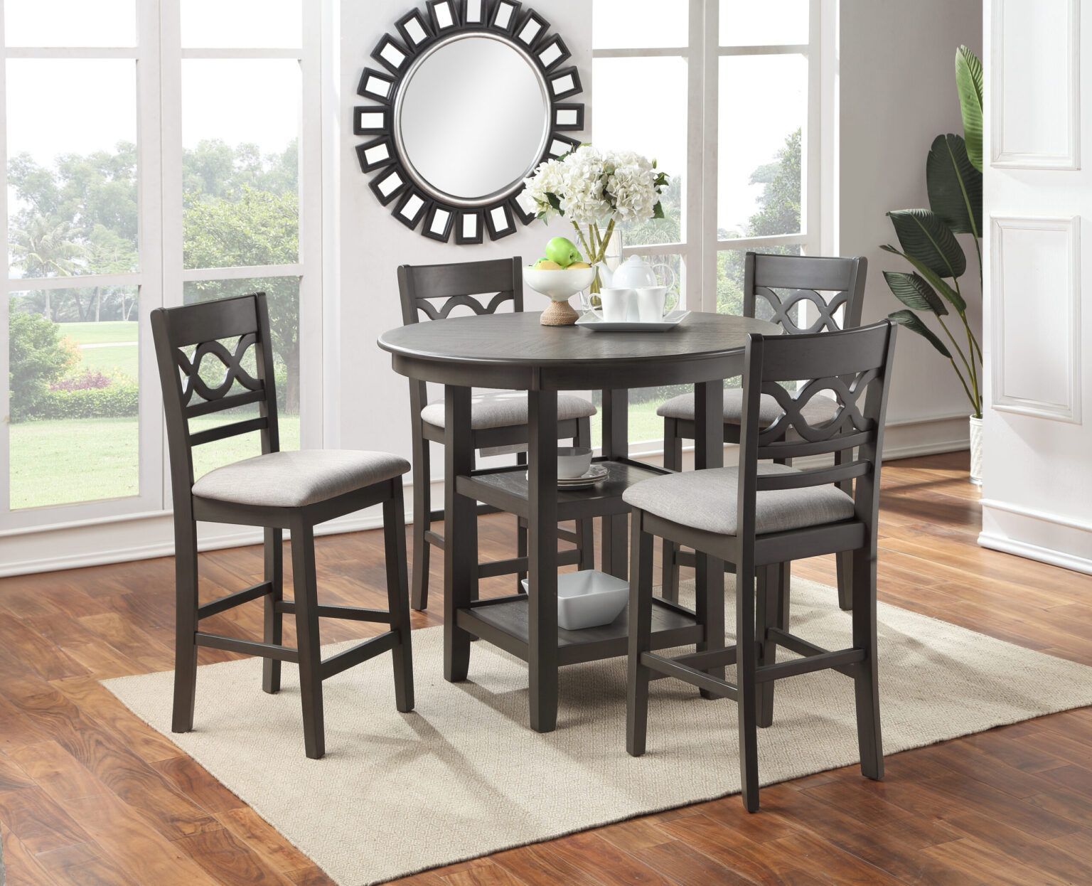 Dark wood dining table set with four chairs on a light rug in a bright room, with a round wall mirror.