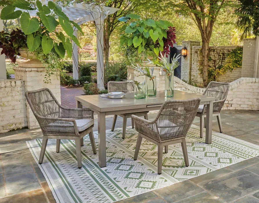 Outdoor patio dining area with a wooden table, woven chairs, and greenery under trees.