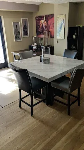 Dining area with a square marble-top table, four black chairs, wall art, and a refrigerator in the background