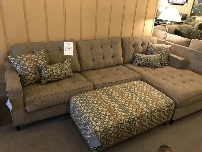 Beige sectional sofa with patterned pillows and ottoman in a showroom setting