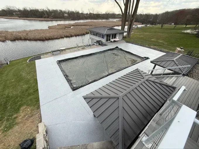 Pool area overlooking a body of water. Gray deck surrounds a covered pool, with gazebo and building nearby.