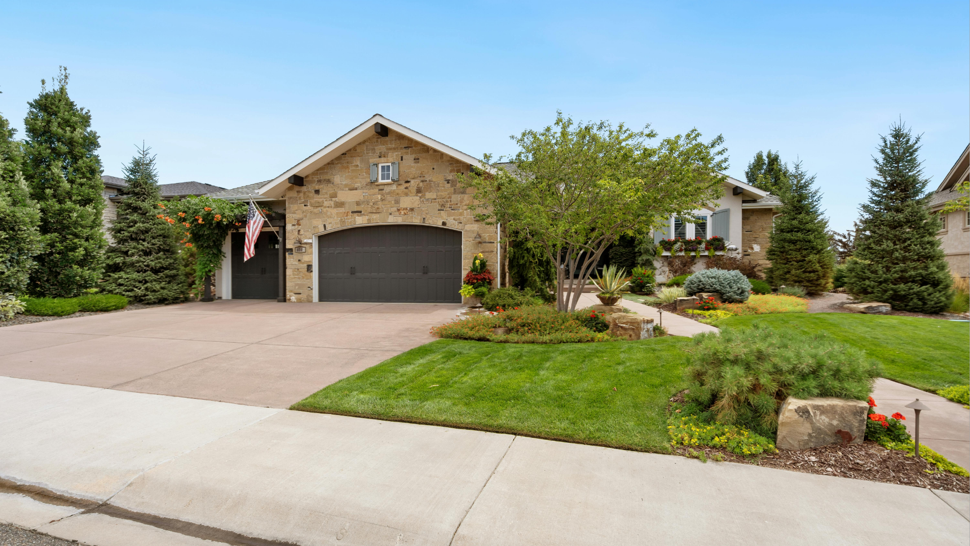 Exterior view of a beige stone house with a two-car garage, lush landscaping, and a blue sky.
