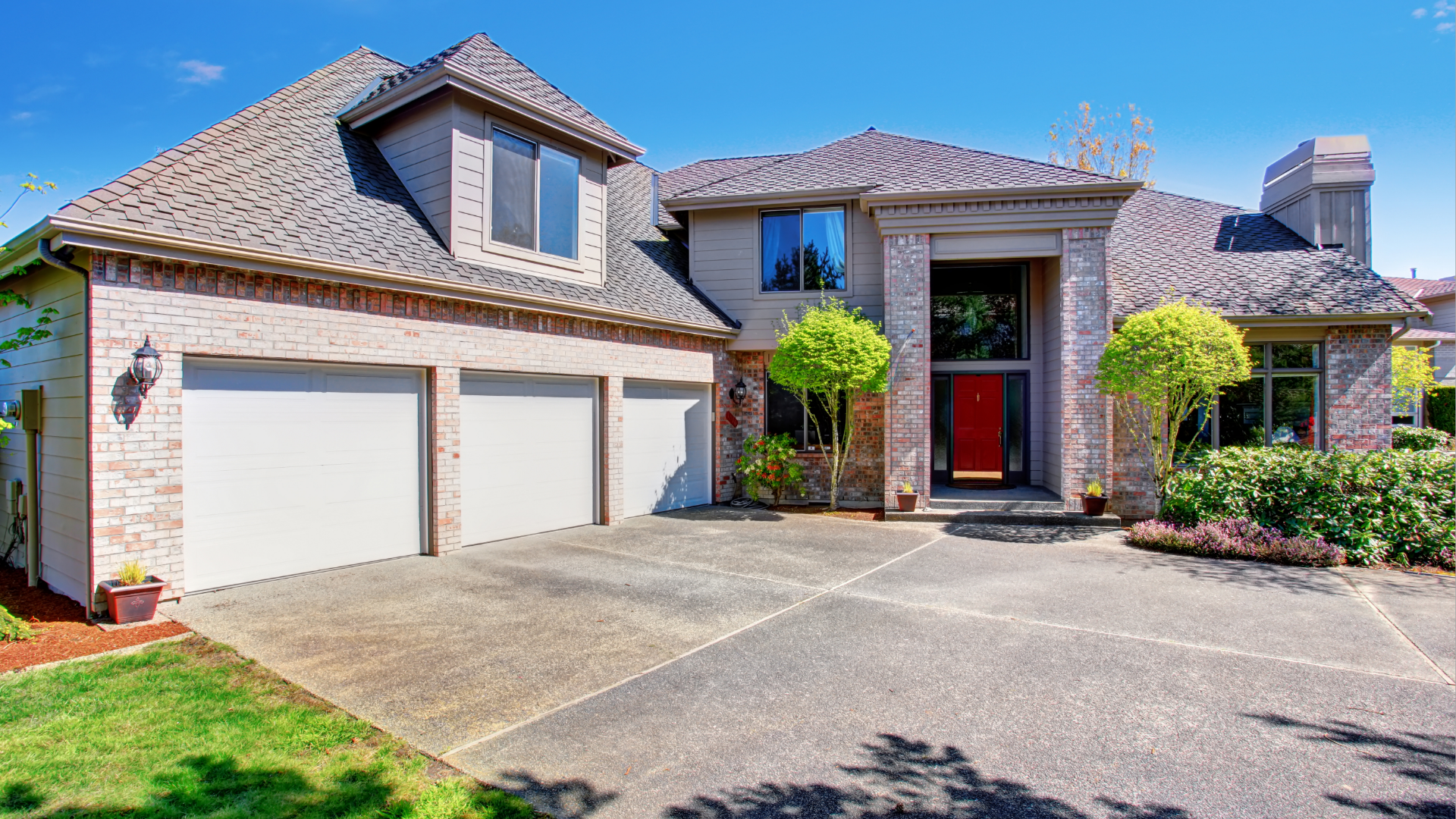 A two-story brick house with three garage doors and a concrete driveway on a sunny day.