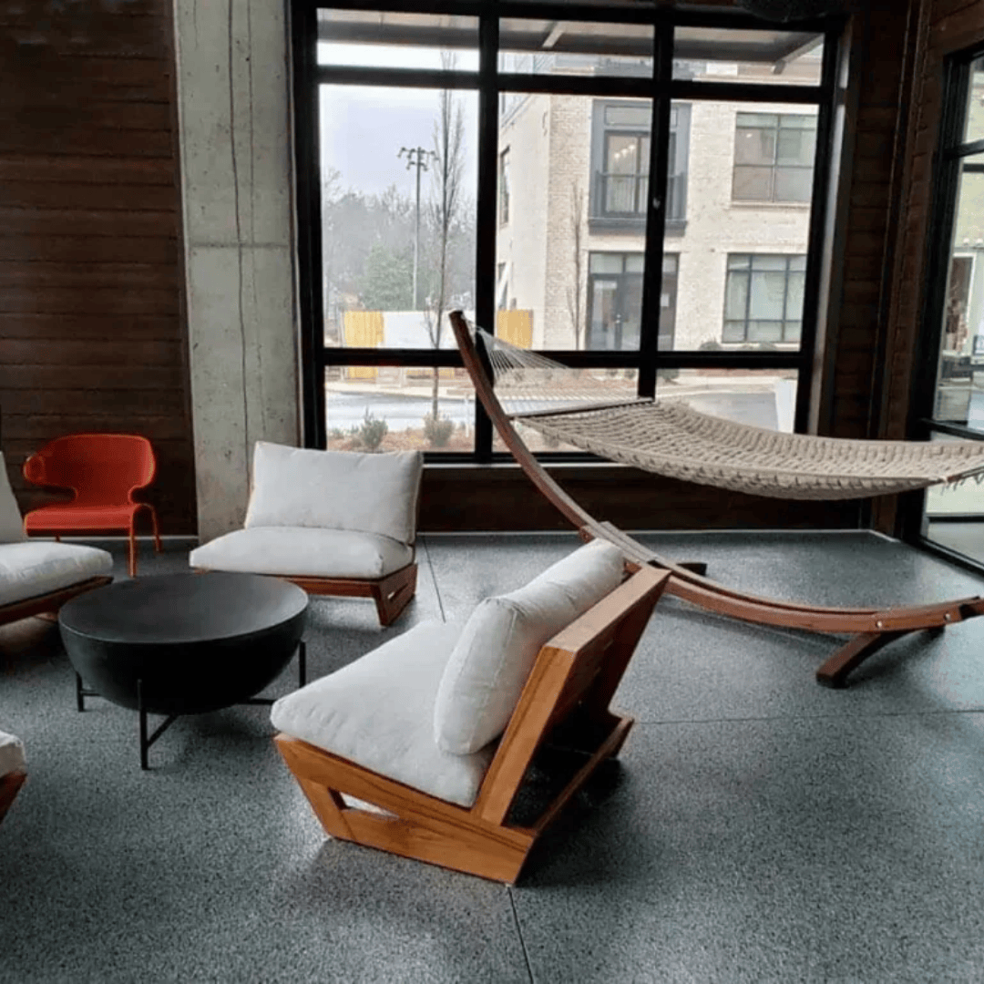 A lounge area with wooden chairs, a hammock, and a dark circular table by a window overlooking a building.