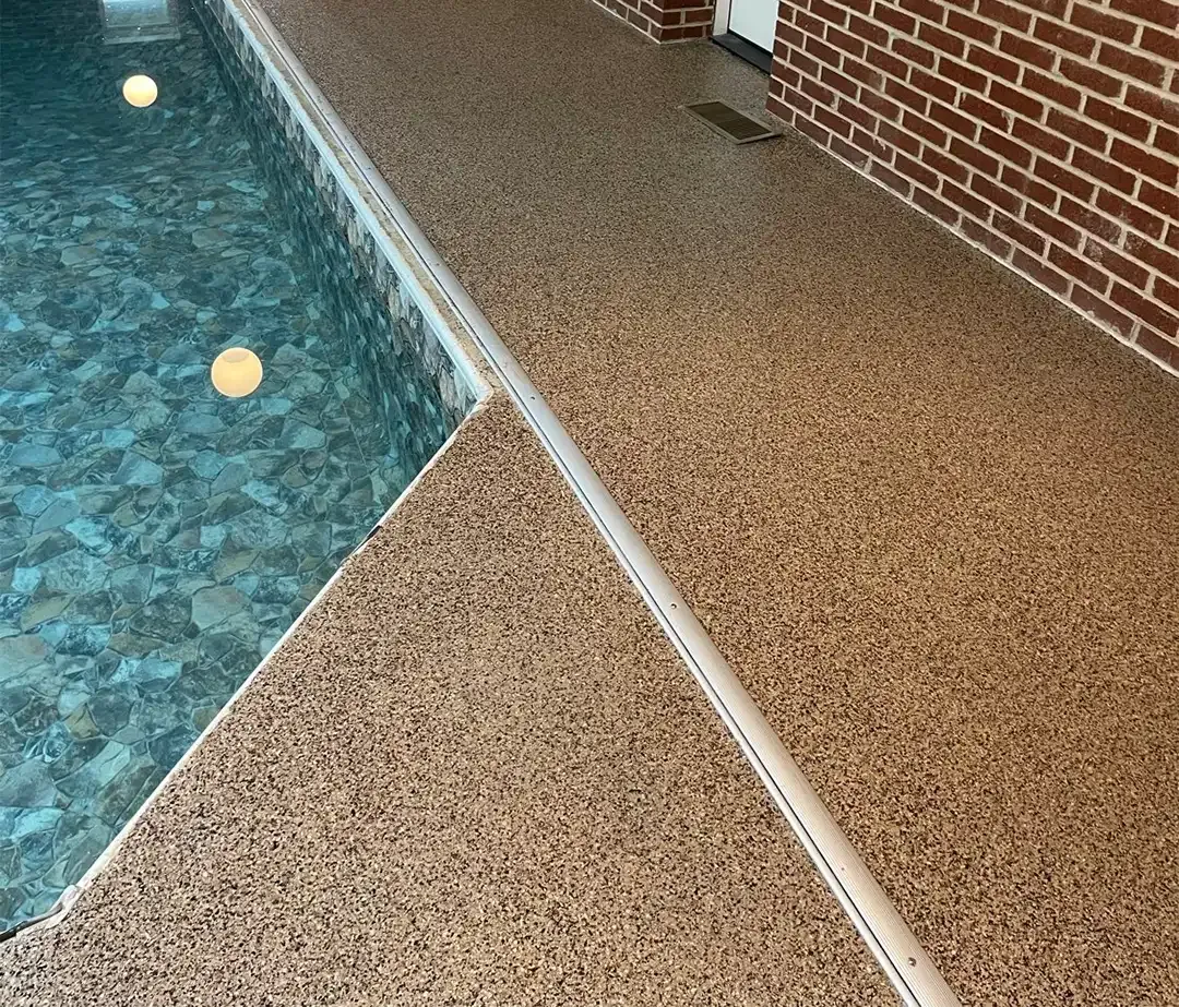 Poolside with textured tan concrete, adjacent to blue water and red brick wall.