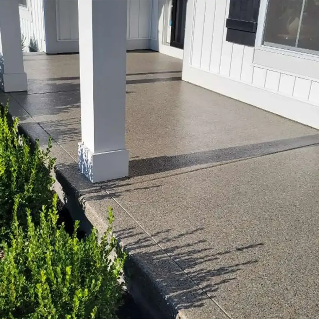 A front porch with a gray speckled concrete surface, white columns, and lush green bushes.