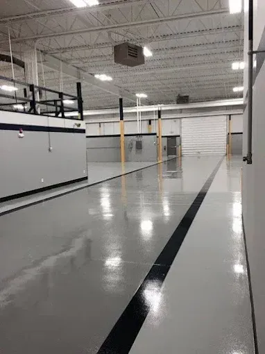 Large, empty warehouse interior with a gray, reflective floor. Black line divides the floor, white walls, and a loading door.