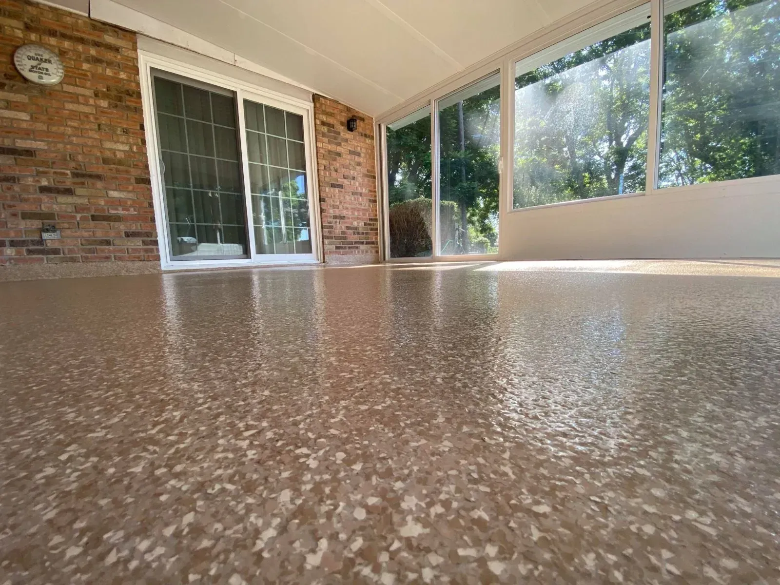 Shiny, speckled concrete floor in a sunroom with brick wall and sliding glass doors. Sunlight streams through windows.