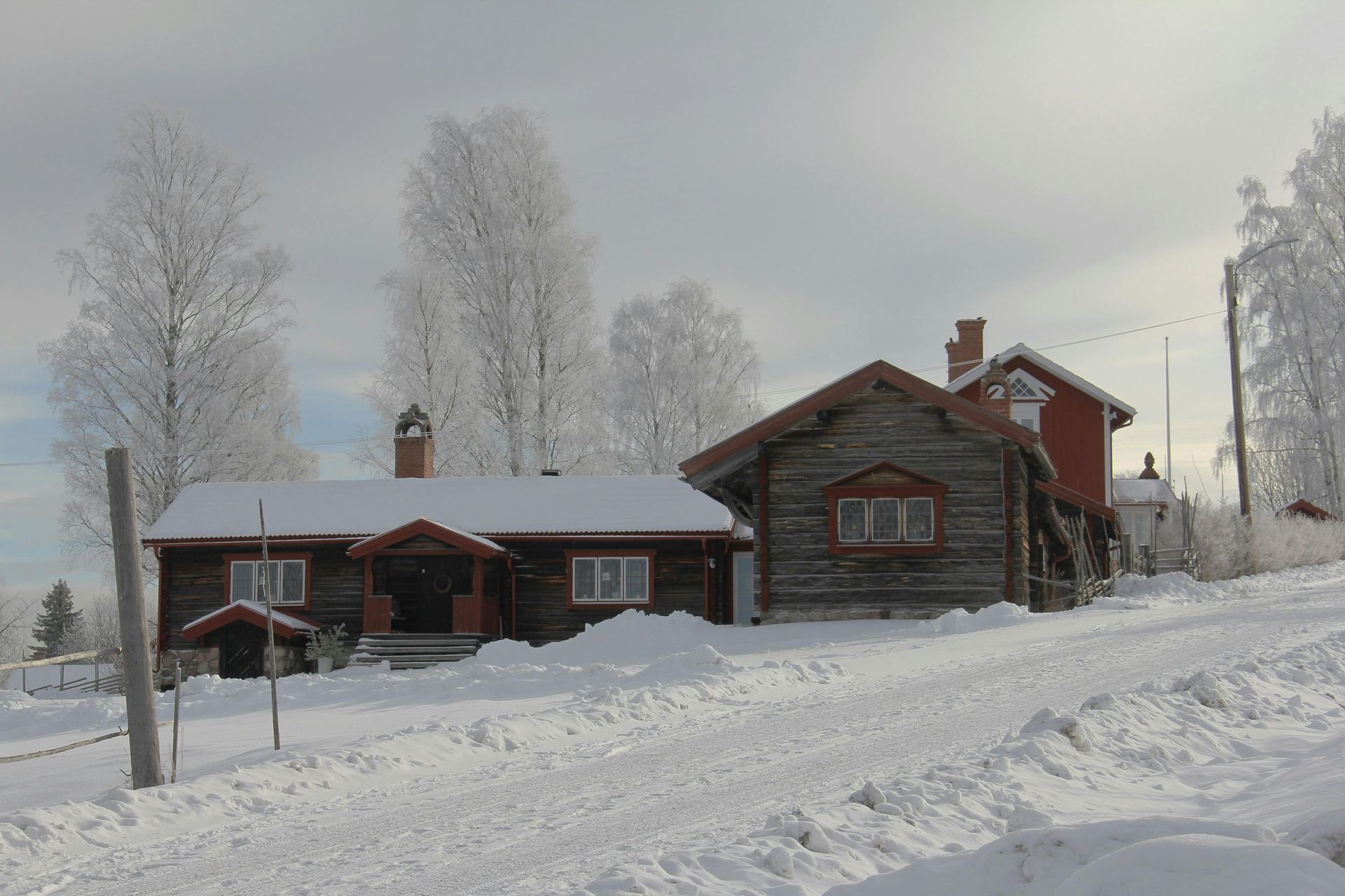 Snow-covered wooden buildings and road in winter landscape. Trees frosted with snow, under a cloudy sky.
