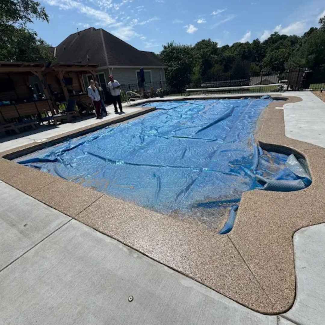 Pool covered with blue tarp, surrounded by brown concrete and people standing. Sunny day.