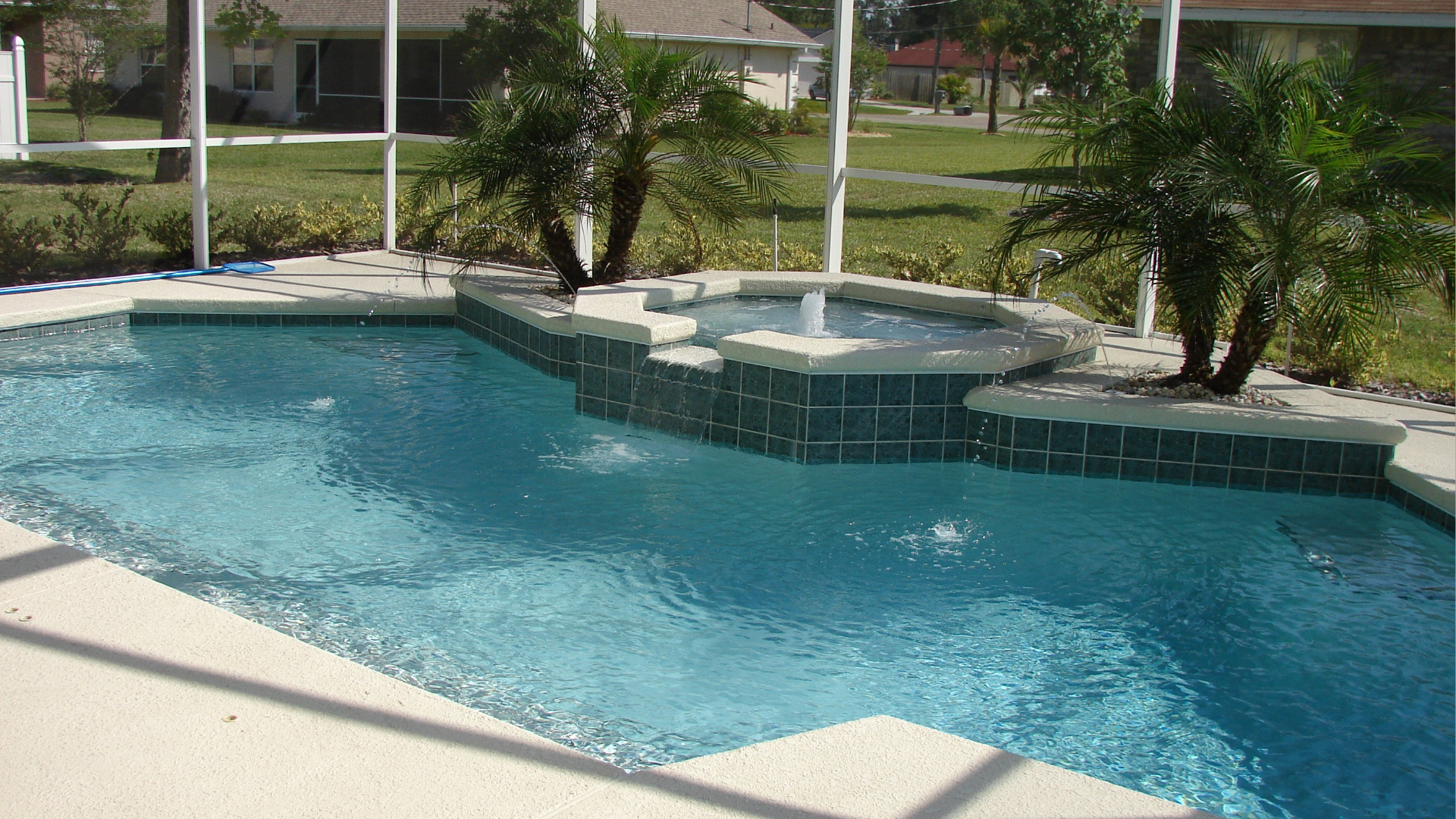 Swimming pool with fountain and palm trees, surrounded by a screened patio and grass lawn.
