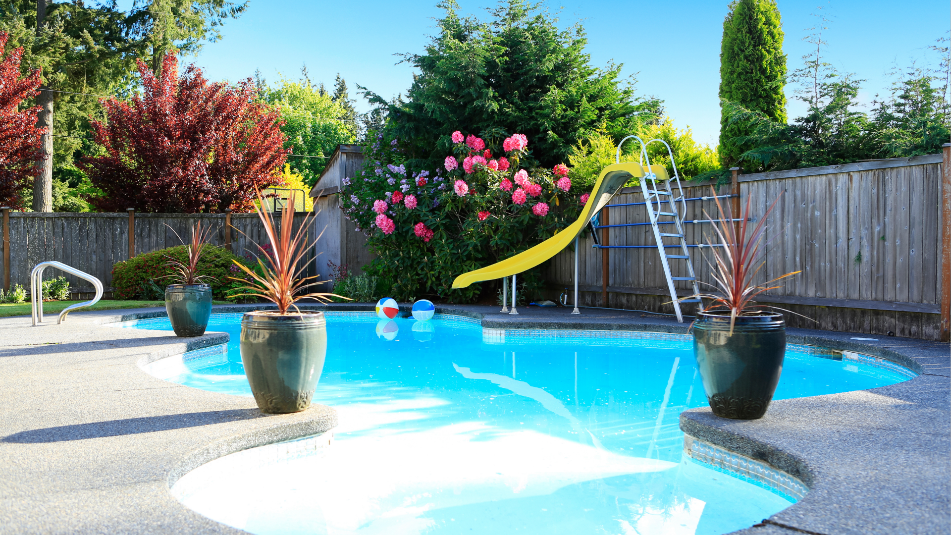 Swimming pool with a yellow slide and ladder, surrounded by a concrete patio and lush greenery.