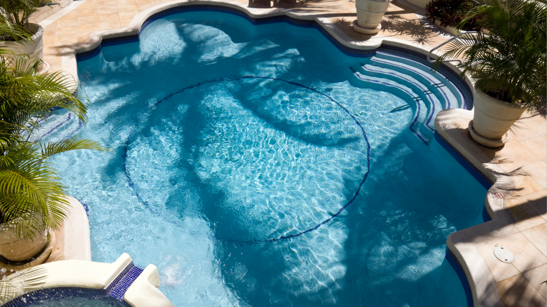 Blue tiled swimming pool with steps, surrounded by stone patio and potted plants.