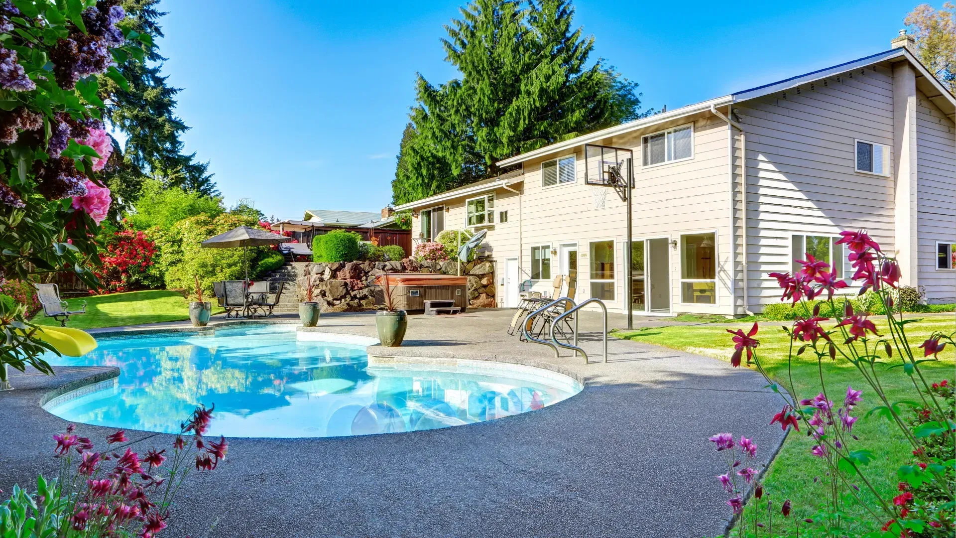 Two-story beige house with a pool, lush green lawn, and flowers under a clear blue sky.