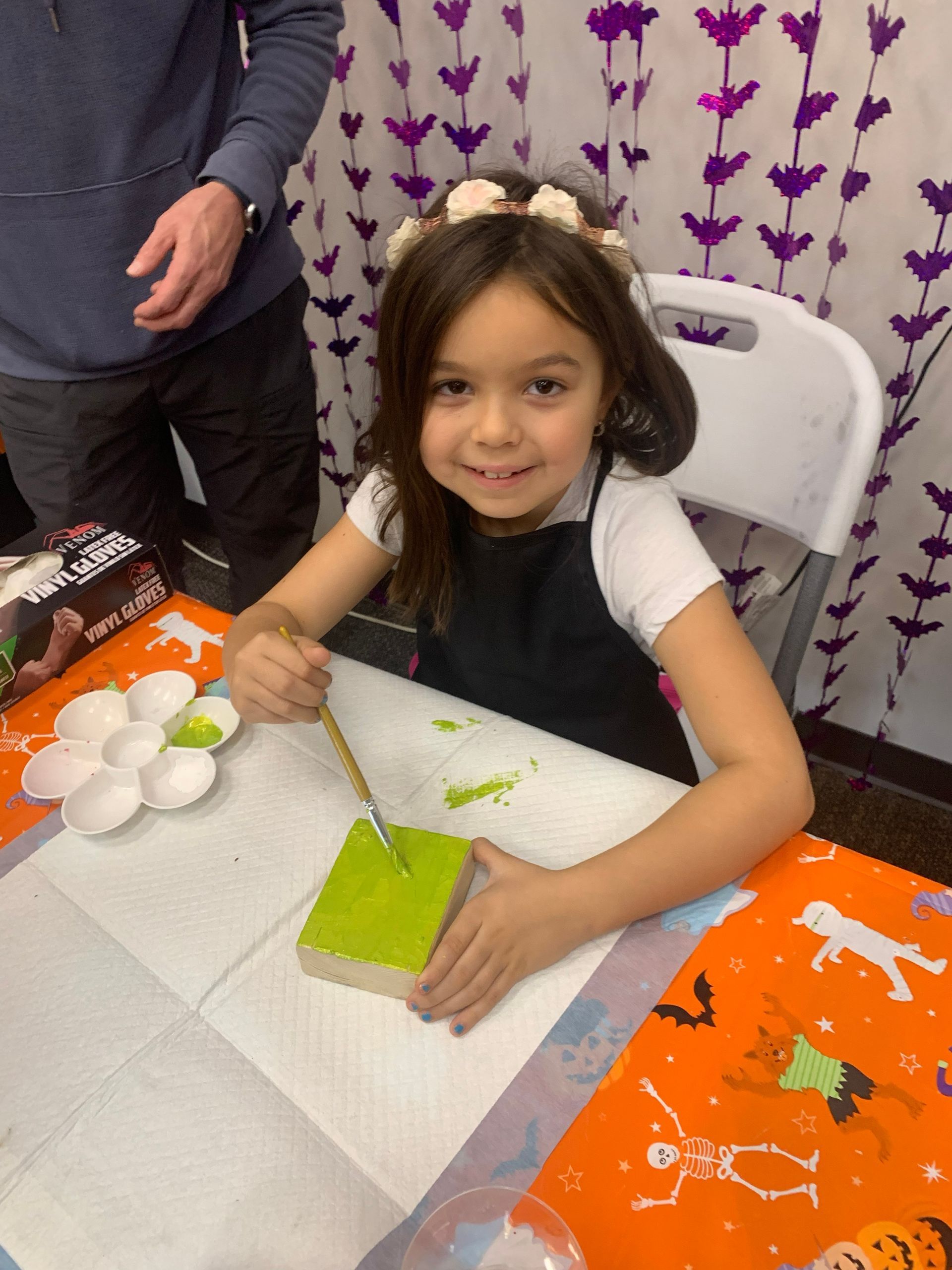 Child smiling at a table, painting a green craft project with brushes and orange supplies nearby.