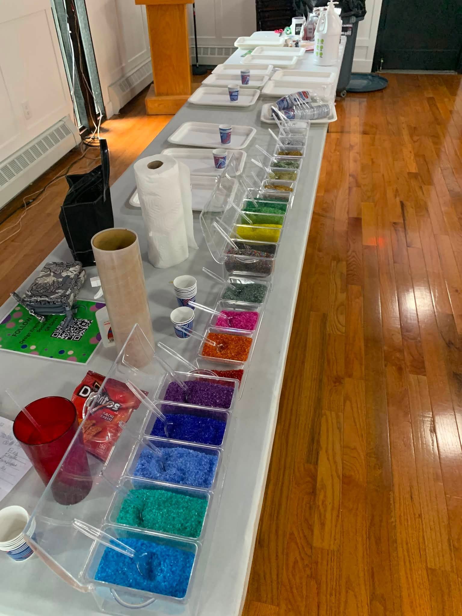 Table of colorful glitter and craft supplies arranged along a room edge
