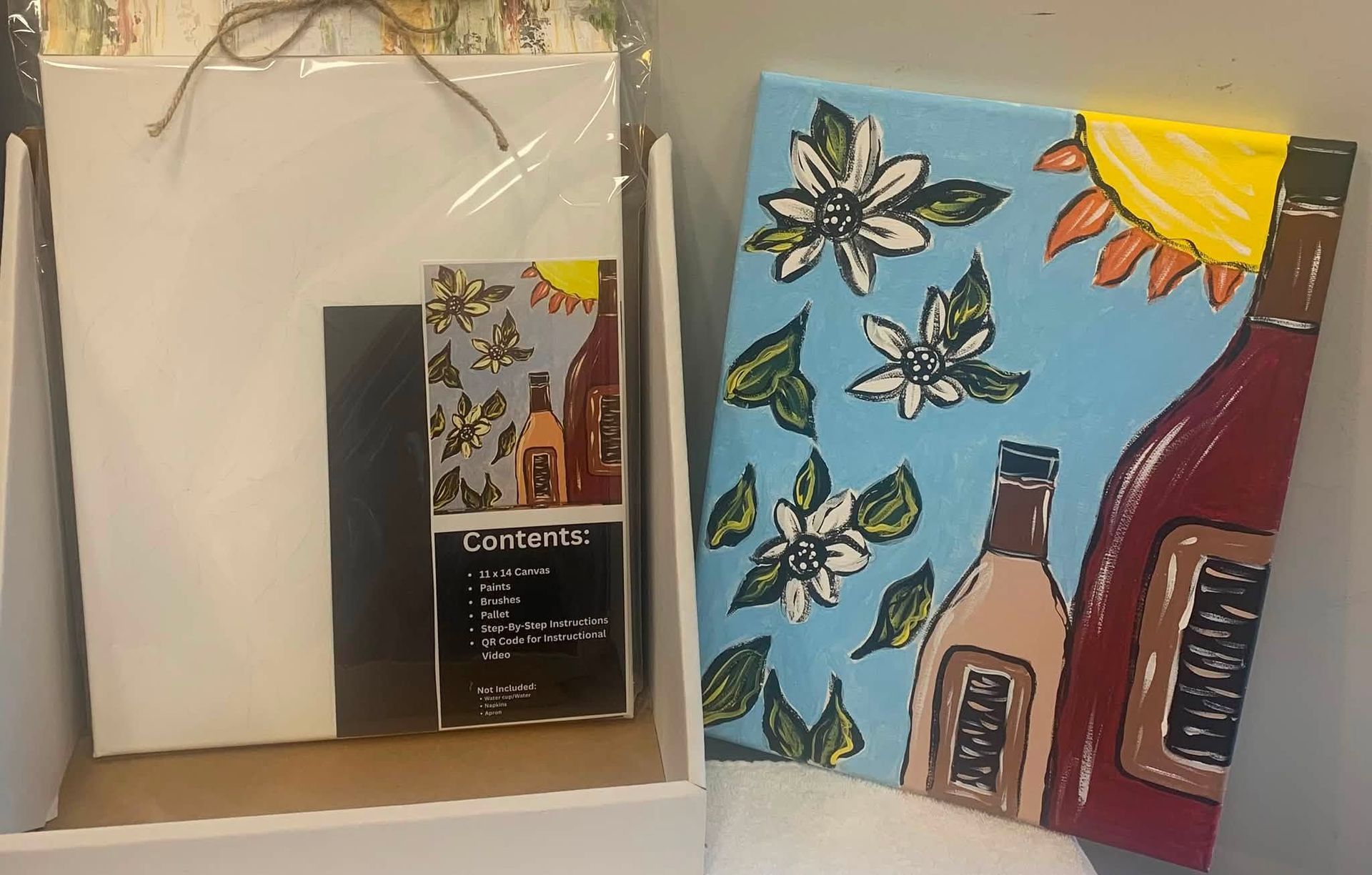 Shelf with two paintings, a blank canvas, and a coffee bag beside a colorful sky-and-bottles artwork.