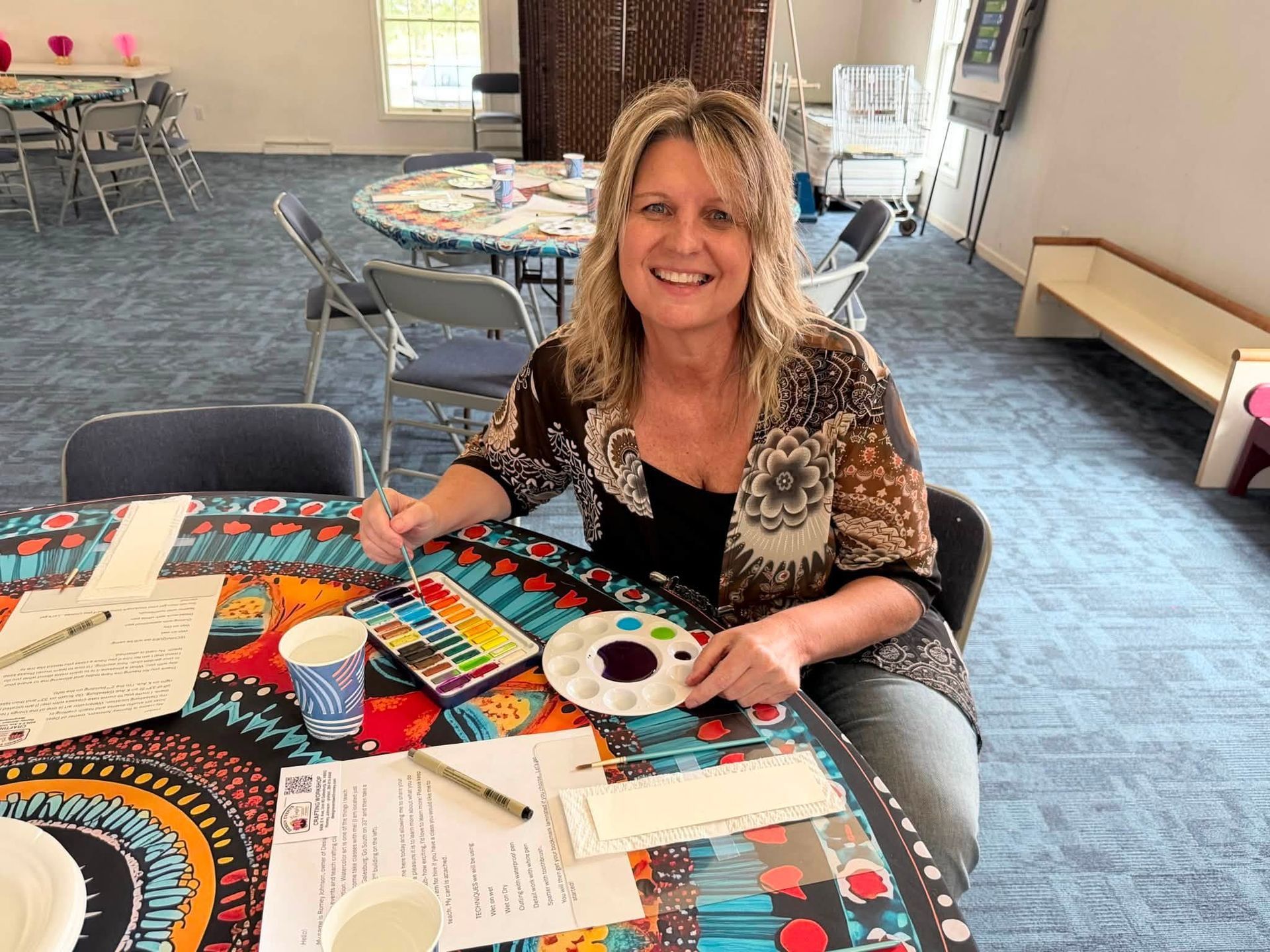 Smiling woman painting at a colorful craft table in a room with chairs and supplies.