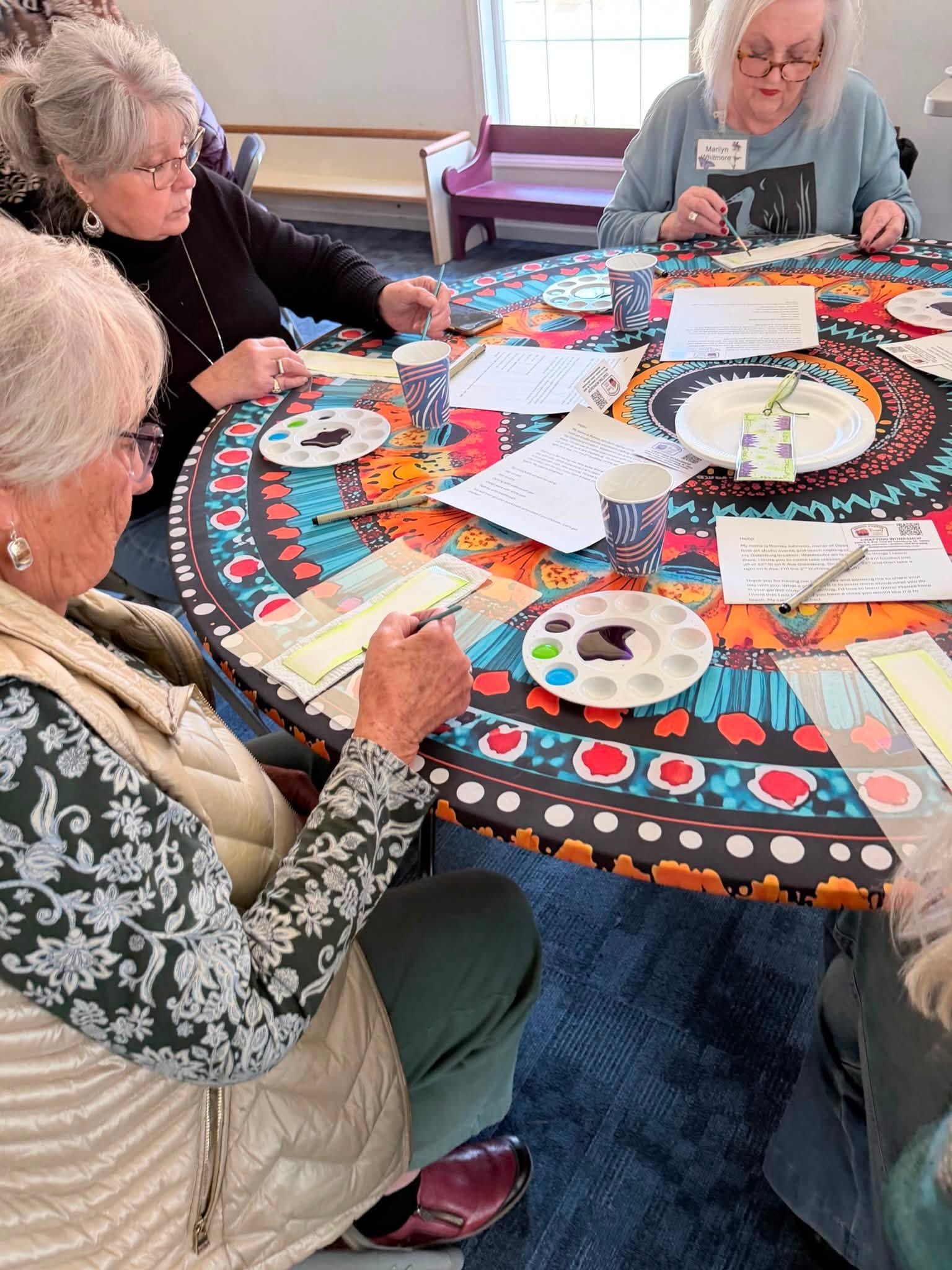Three people paint a colorful circular canvas at a table with brushes and palettes.