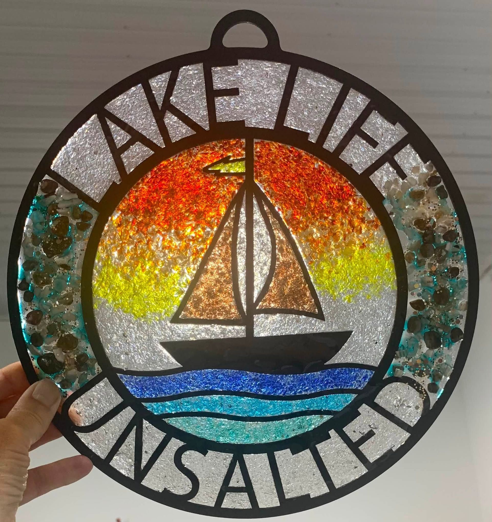 Circular stained-glass-style sign with a sailboat over a sunset, reading “Lake Life Wisconsin.”