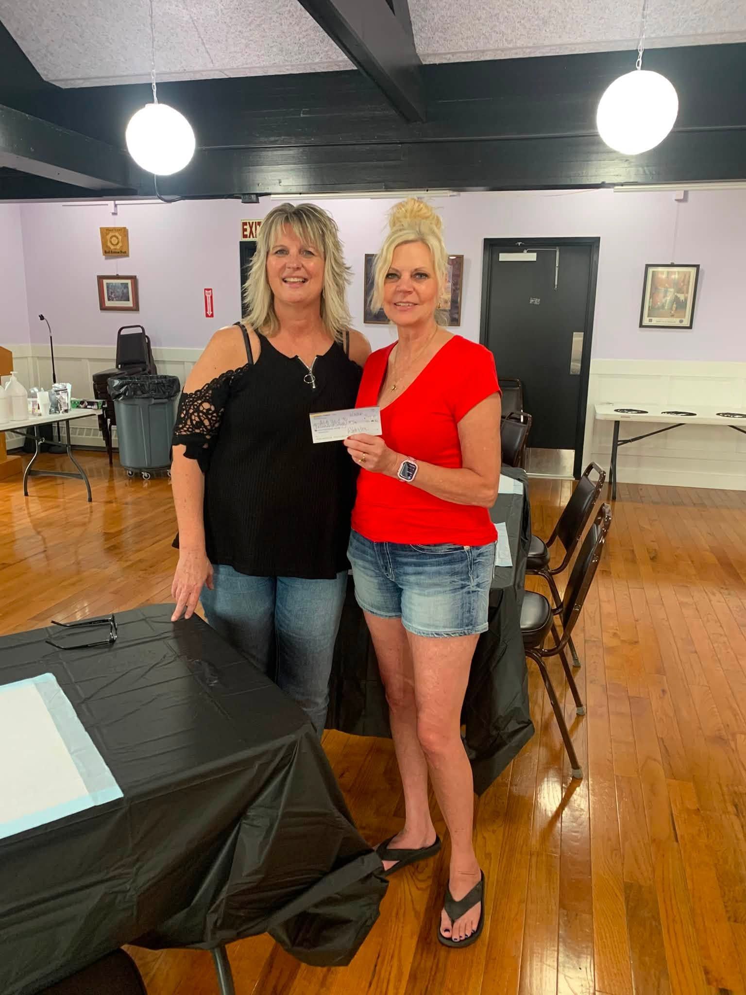 Two women standing indoors, one holding a check, in a community hall with tables and chairs
