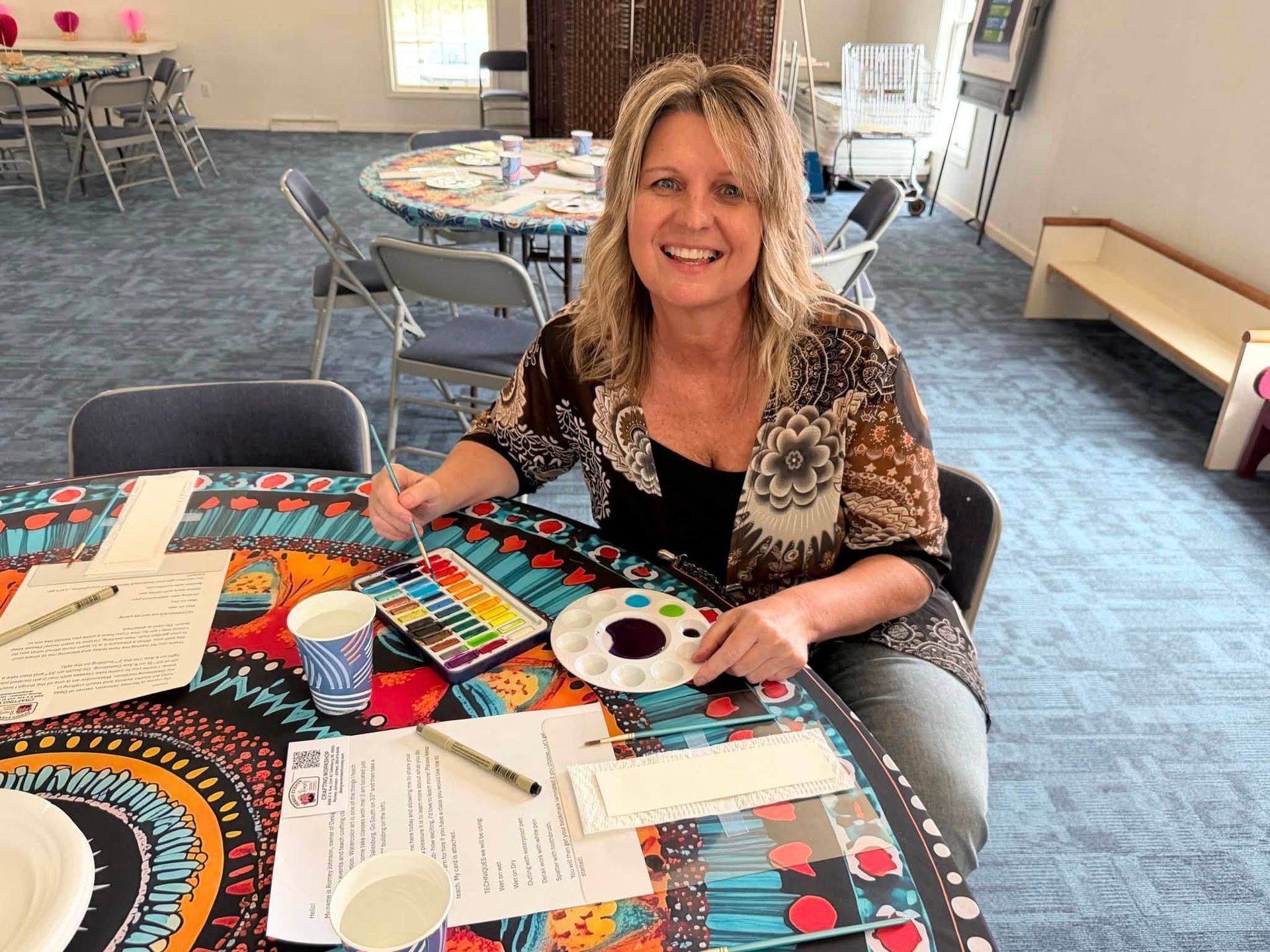 Woman at a craft table holding a paint palette in a bright community room