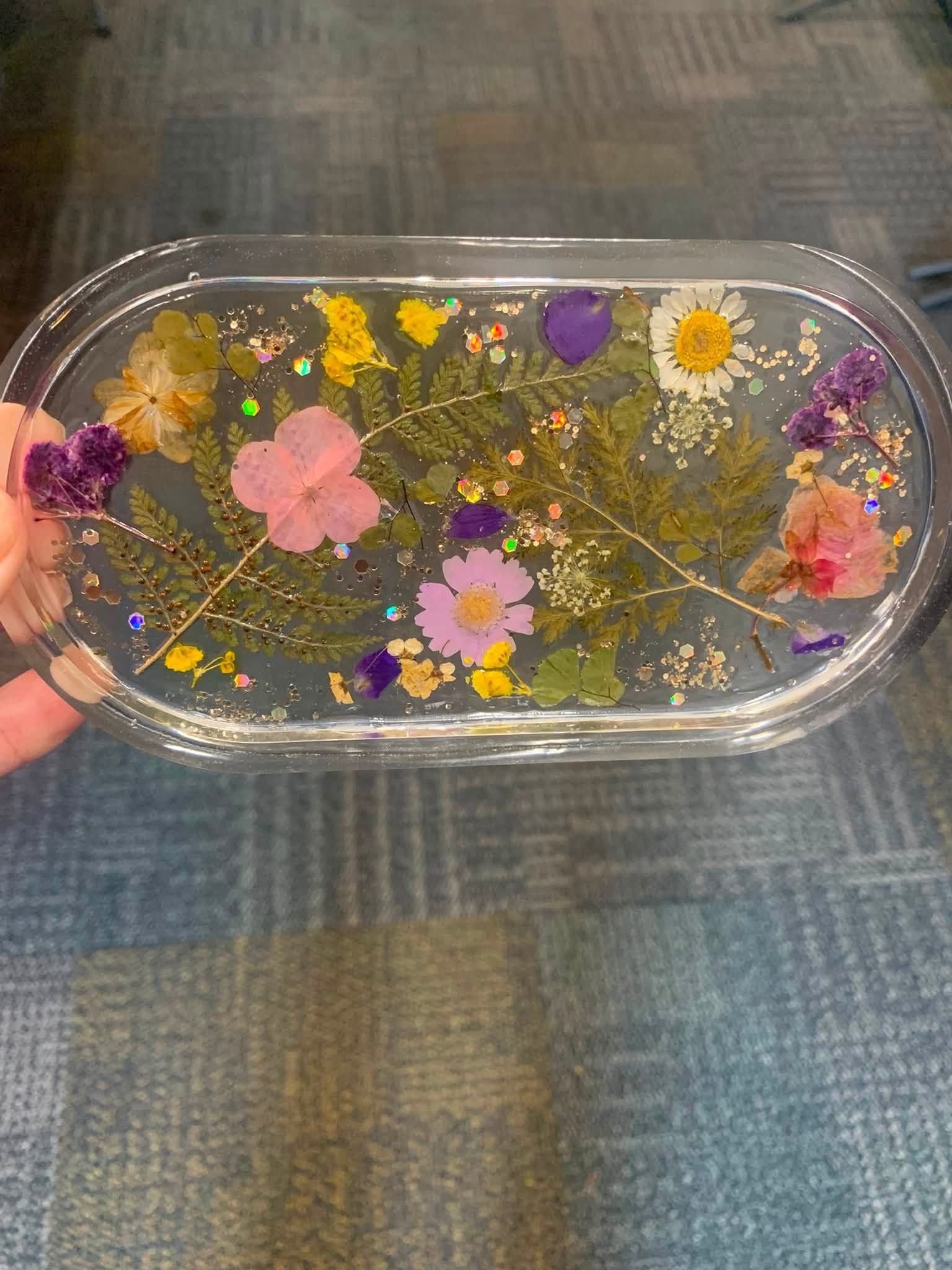 Clear oval tray with floating dried flowers and gold flakes on a gray fabric background