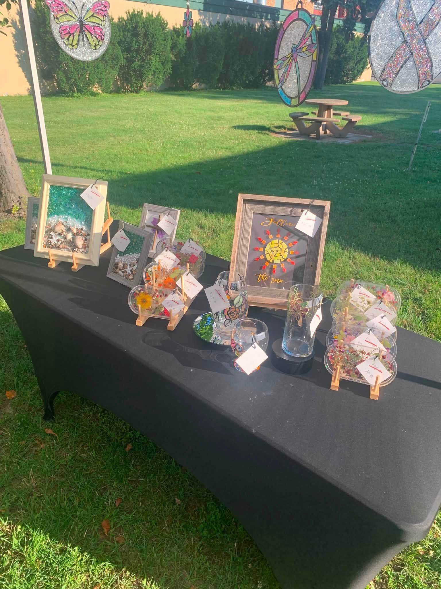 Small craft display on a black table outdoors, with framed floral artworks and decorative figurines on grass.
