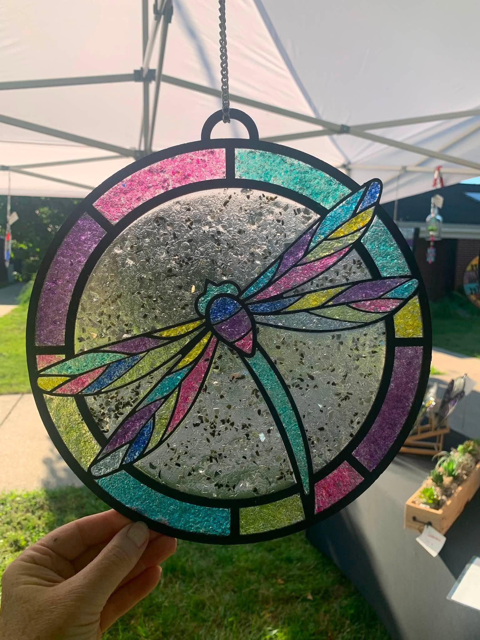 Colorful circular stained-glass dragonfly ornament held up outdoors under a canopy