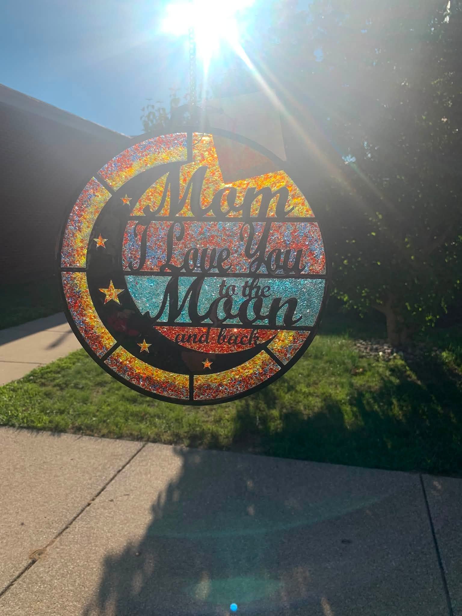 Colorful circular “Mum” Mother’s Day yard sign on a sunny lawn by a sidewalk.