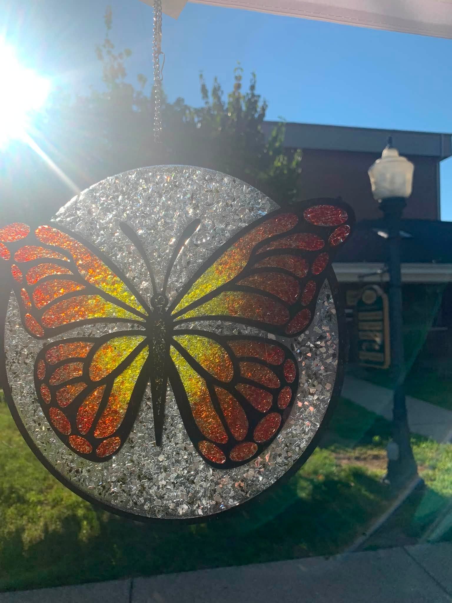 Colorful butterfly mosaic hanging outdoors in sunlight, with orange, yellow, and red wings on a silver circle.