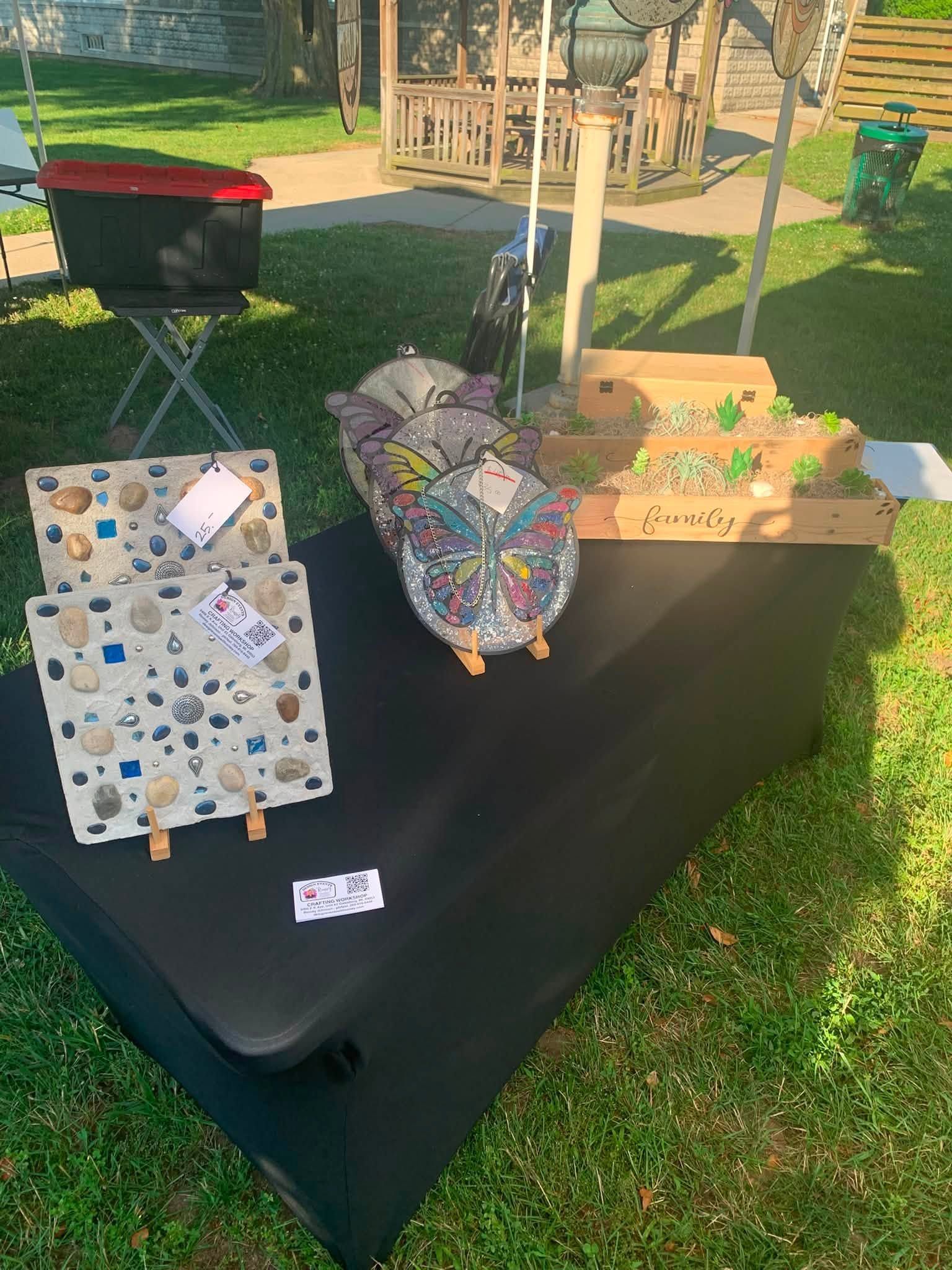 Craft fair display table with mosaic garden art, colorful decorated items, and a small sign outdoors on grass