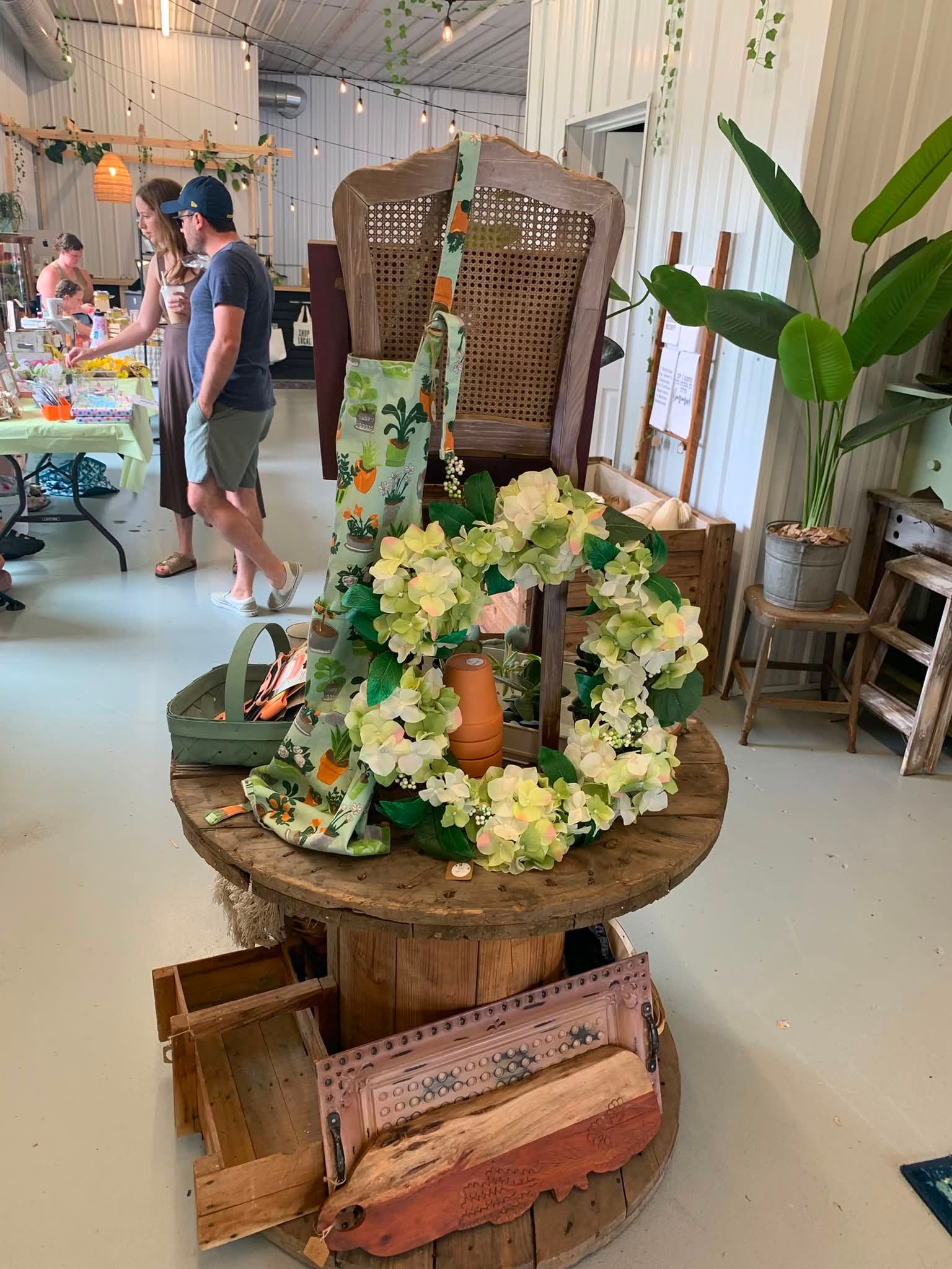 Flower arrangement on a round wooden table in a bright indoor market or café, with a tall decorative chair behind it.