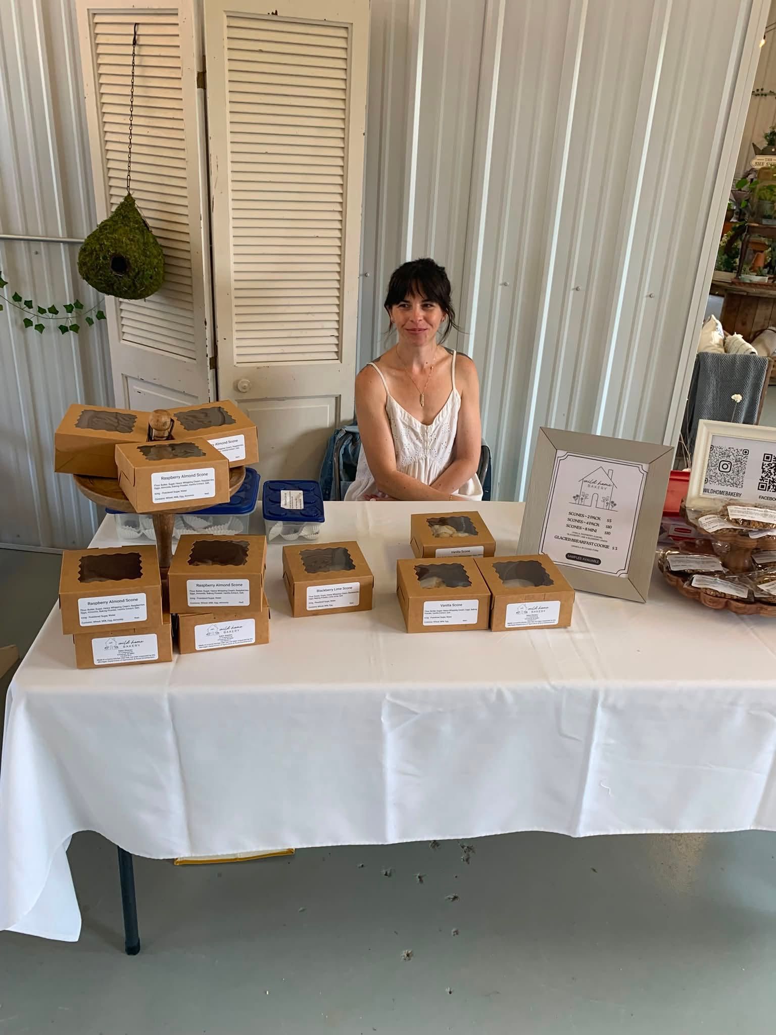 Vendor booth with labeled boxes of baked goods on a white table and a seated woman behind it