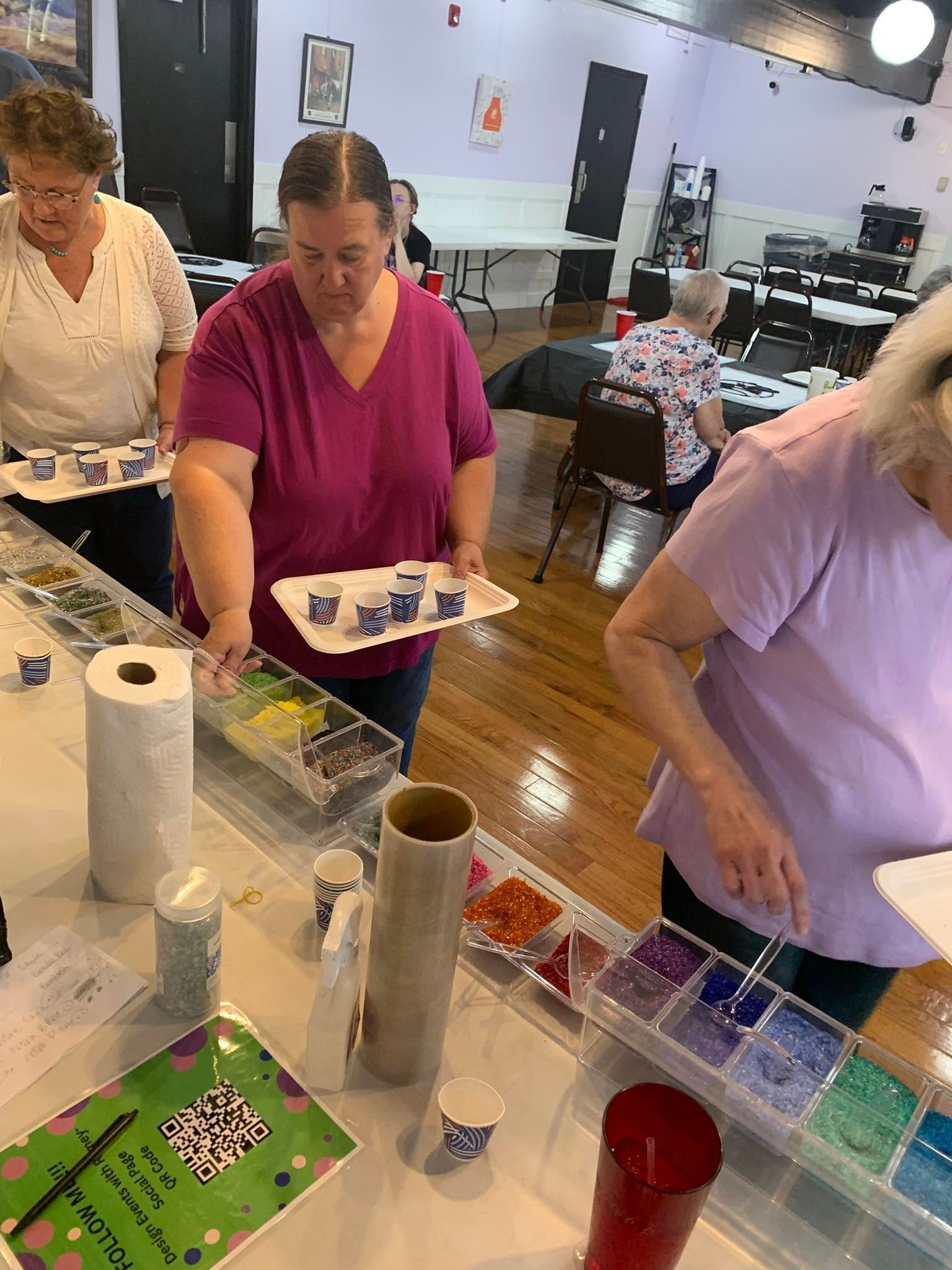 People serving snacks at a community table with cups, bins, and paper towels in a bright room