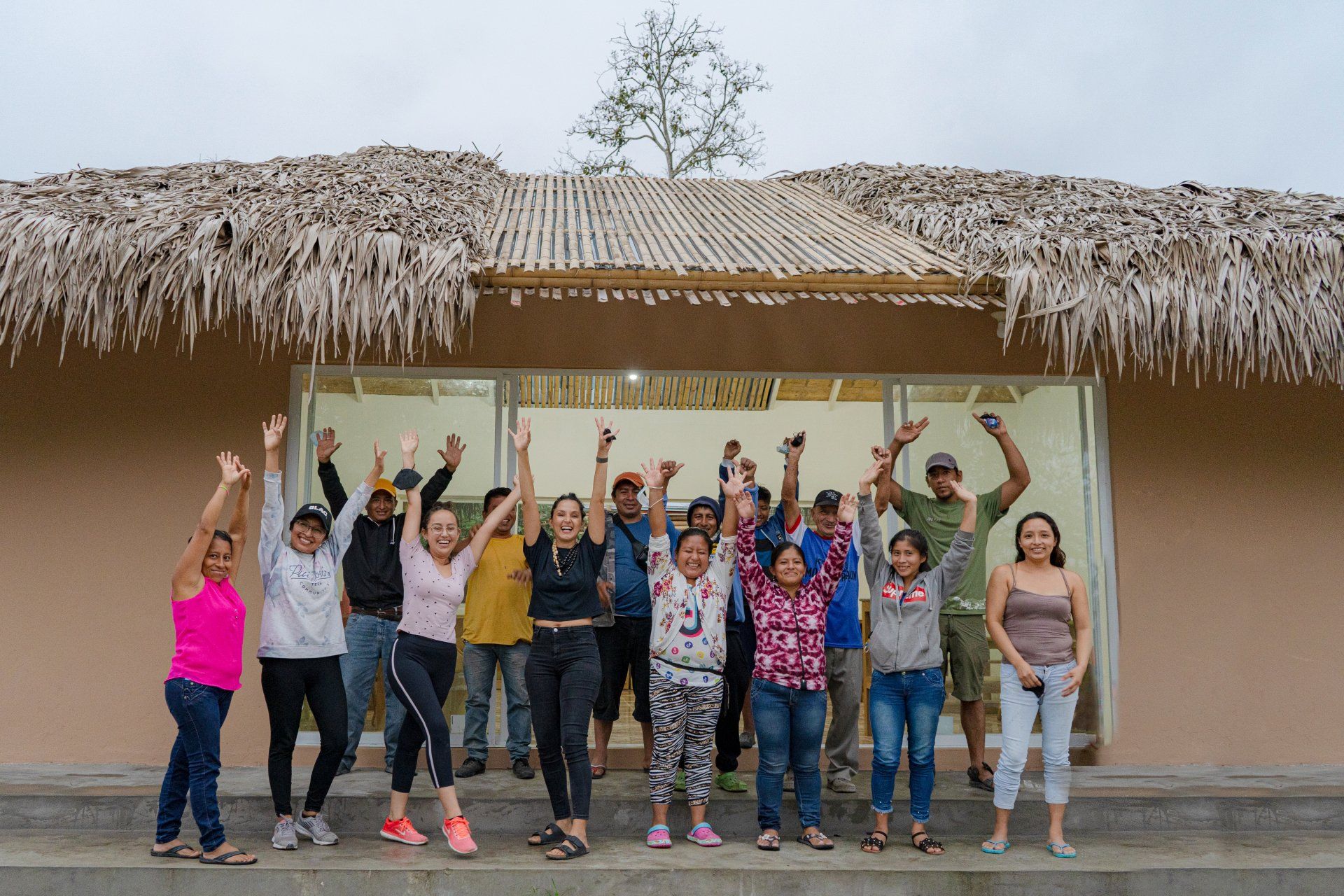 Group of people with arms raised, in front of a building with a thatched roof.