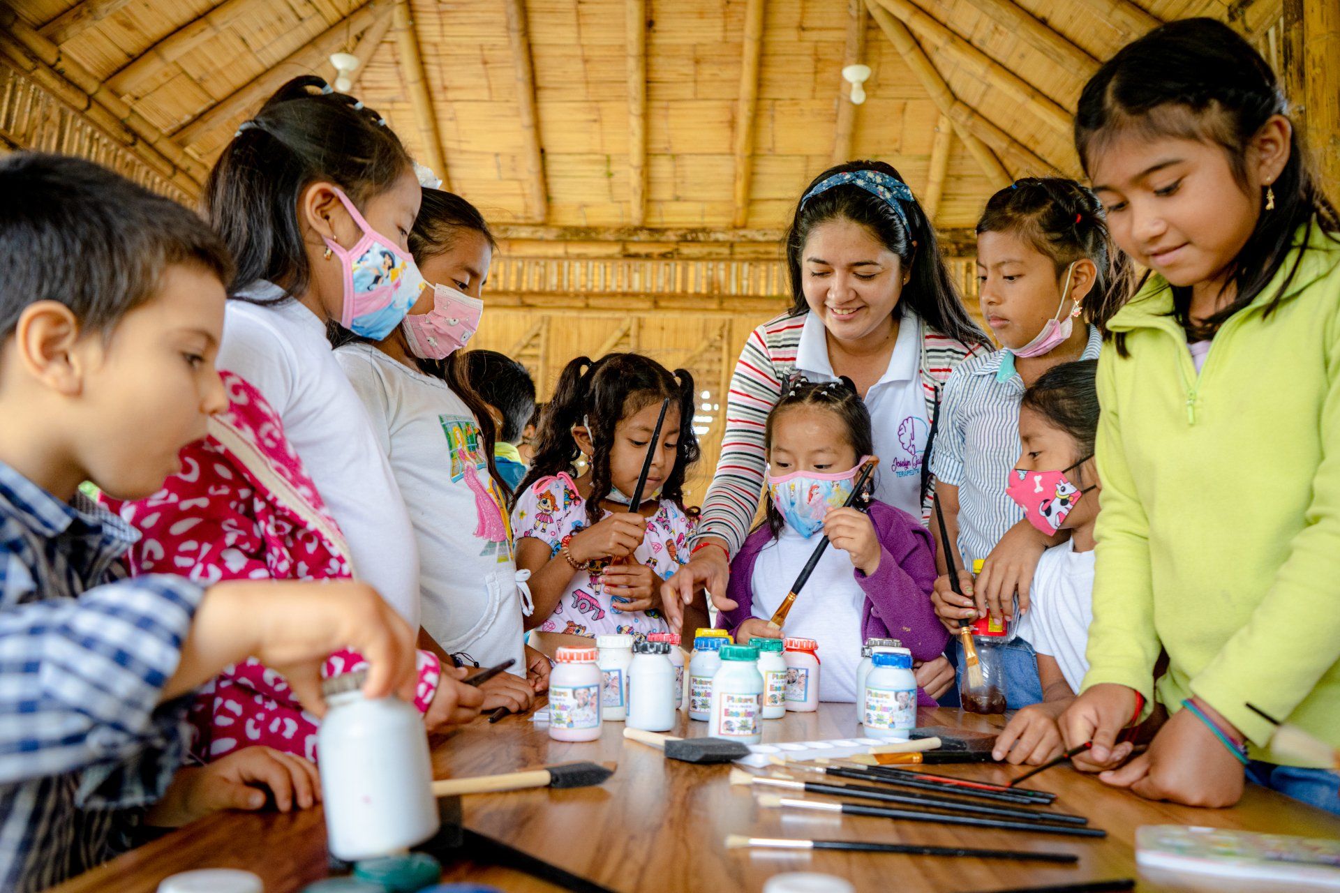 A group of children and a woman crafting at a table, some wearing masks, in a bamboo structure.