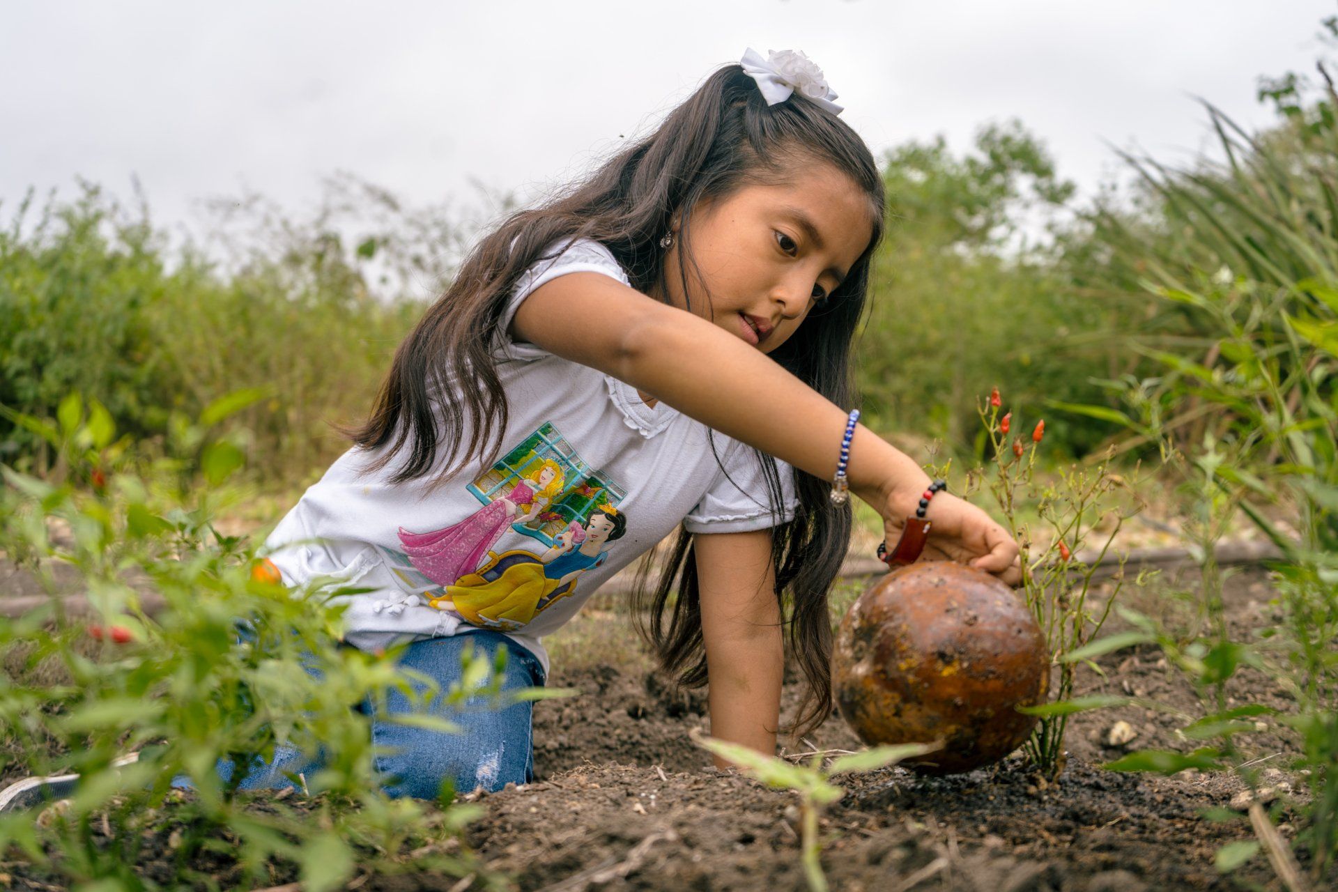 Girl with long hair, in a garden, examines a round object. She wears a white shirt, jeans, and a flower in her hair.