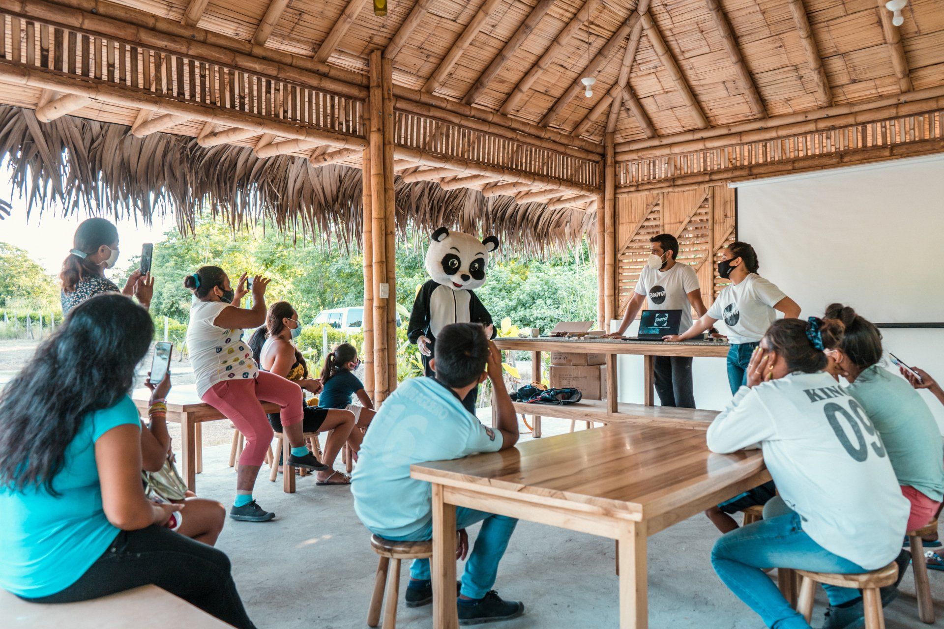 People in a bamboo hut, watching a presentation. A panda mascot stands with a presenter.