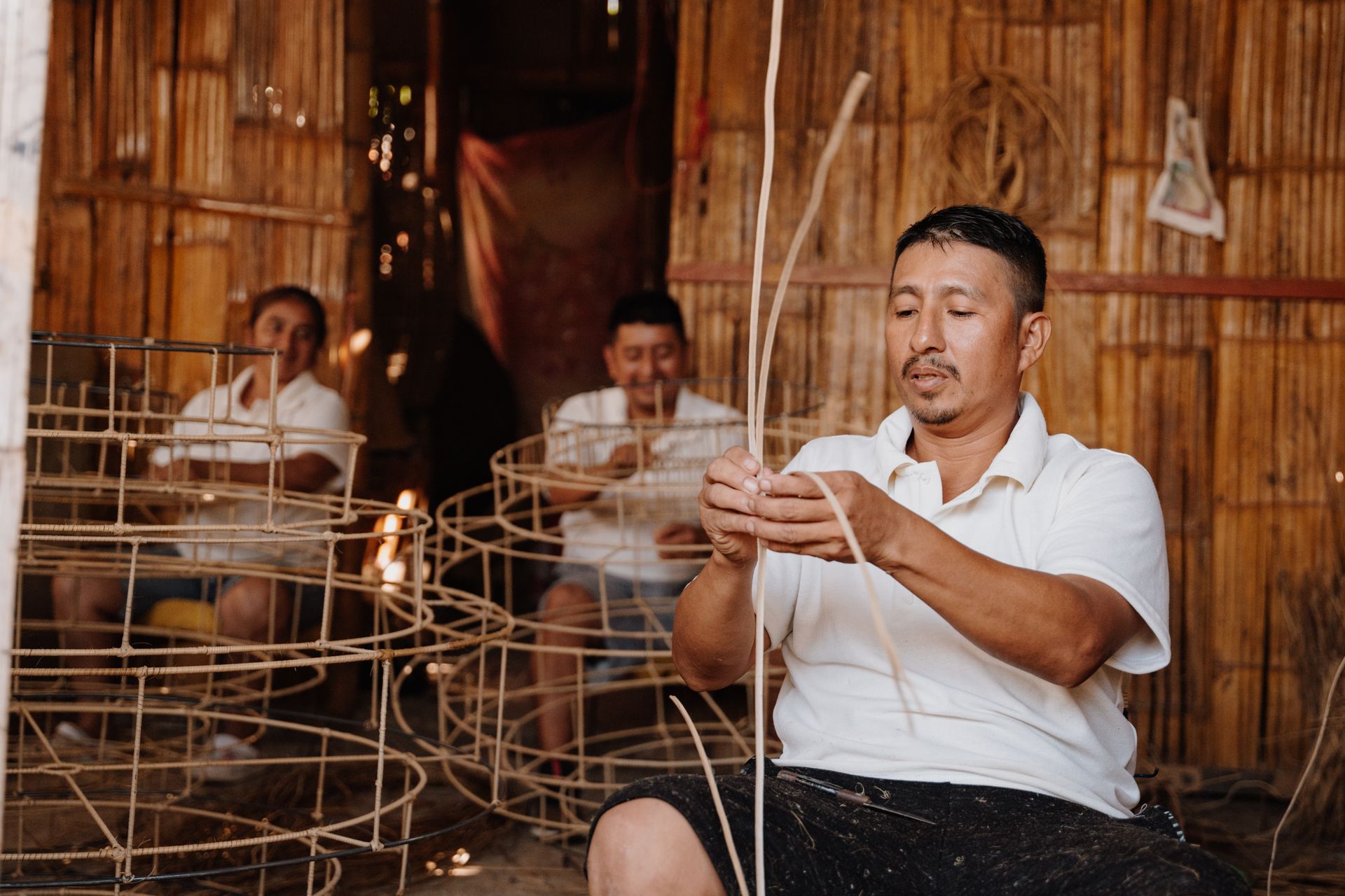 Man weaving basket with two others in a workshop; wooden frames, brown weave, light clothing.