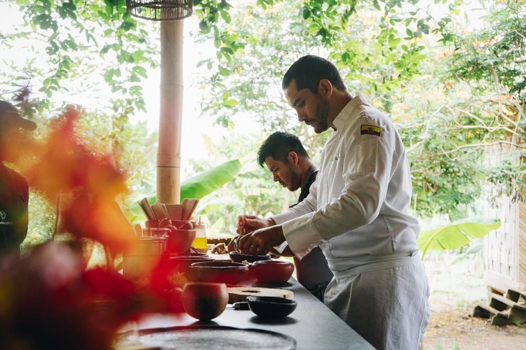 Two chefs preparing food in an outdoor kitchen with greenery in the background.