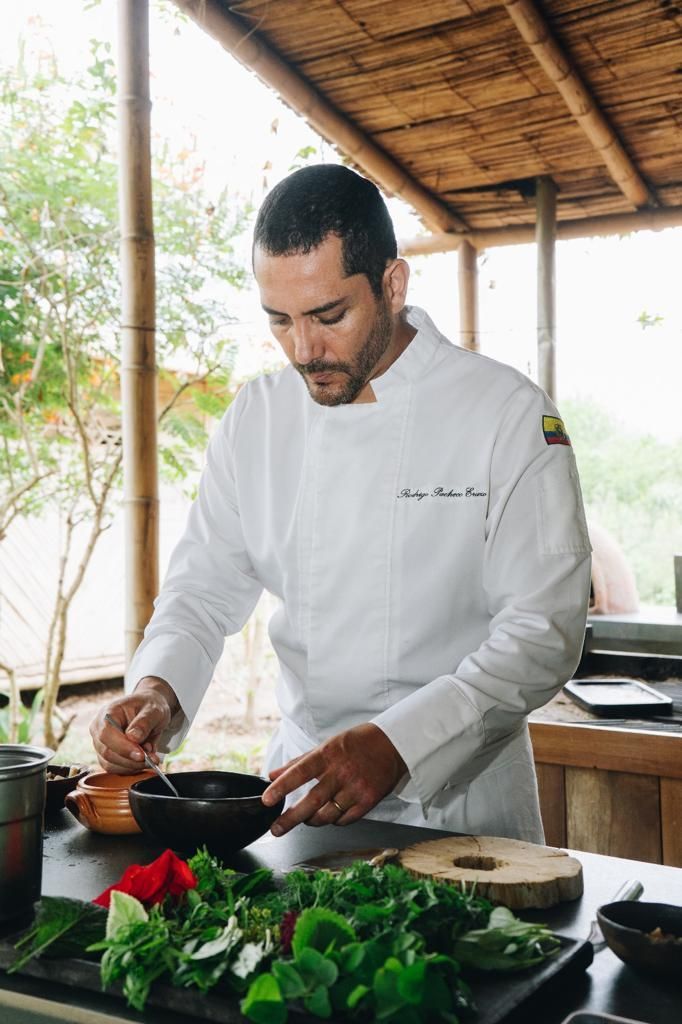 Chef in white coat preparing food outdoors, near plants and wooden surfaces.